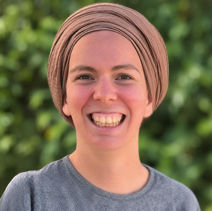 תמונת פרופיל – ימימה שינדלר, מאמנת אישית תמונת דיוקן של ימימה שינדלר, מחייכת ועוטה כיסוי ראש בצבע בז׳, על רקע צמחייה ירוקה.